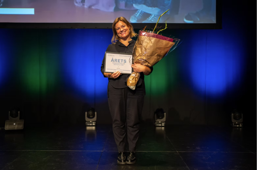 Kvinde der står med en buket blomster og et billede, hvor der står 'årets demensvenlige initiativ'
